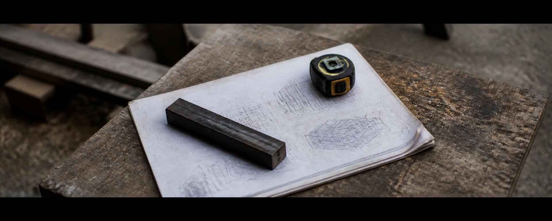 Printed engineering documentation lying open on a dusty site bench, weighted down by a steel offcut